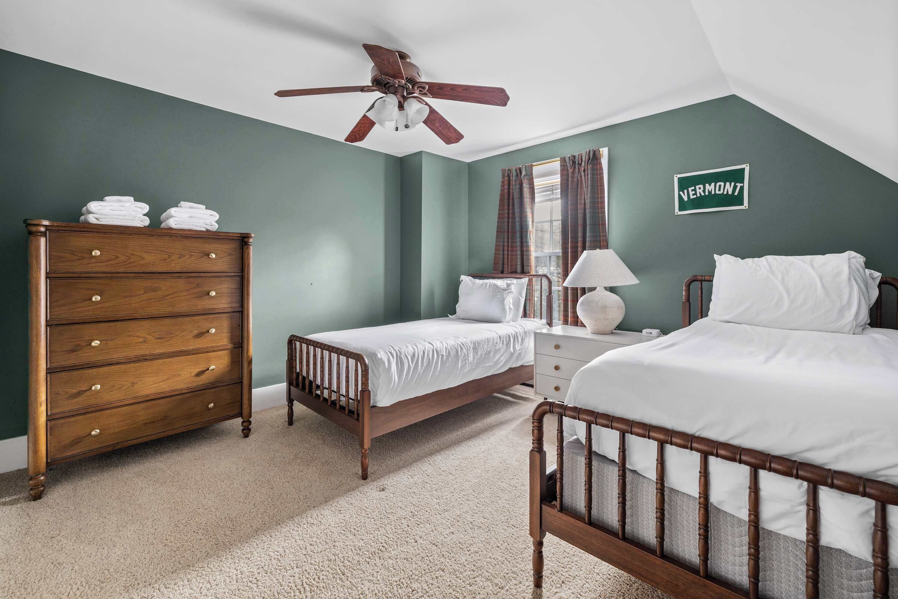 A bedroom with a wooden dresser, two beds, a nightstand, a lamp, a ceiling fan, and a Vermont sign.