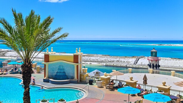 Área de piscina de um resort com vista para o mar, uma palmeira e um pequeno edifício com uma fonte.