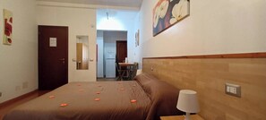 Ein Hotelzimmer mit Bett, Nachttisch, Lampe, einer Tür und Blick in den Küchenbereich.