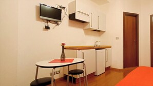 A compact room with a kitchenette, a single bed, a TV mounted on the wall, and a door.