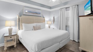 Ein ordentlich eingerichtetes Schlafzimmer mit Bett, Nachttischlampen, Deckenventilator, einem Gemälde und einem Fenster mit Vorhängen.