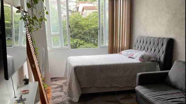 A bedroom with a bed, a sofa, a TV, a plant, and a window with a view of greenery. - Charming 1-bedroom loft in downtown Rio de Janeiro. (Rio de Janeiro)
