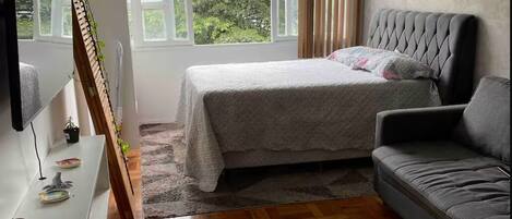 Un dormitorio con cama, sofá, televisión, planta y ventana con vistas a zonas verdes.