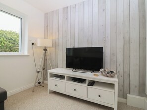 A modern living room with a flat-screen TV on a white TV stand, a lamp, and a window with a view of greenery. - Red Wharf Retreat (Pentraeth)