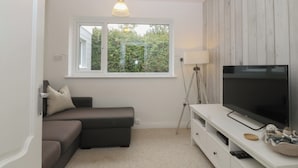 A room with a sectional sofa, a flat-screen TV on a white stand, a window with a view of greenery, and a floor lamp. - Red Wharf Retreat (Pentraeth)
