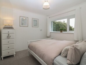 A bedroom with a bed, a dresser, two framed pictures, a window with curtains, and a hanging light fixture. - Red Wharf Retreat (Pentraeth)