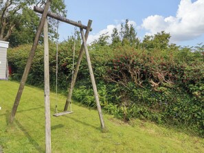 A wooden swing set in a garden with lush greenery and a bush with red berries. - Red Wharf Retreat (Pentraeth)