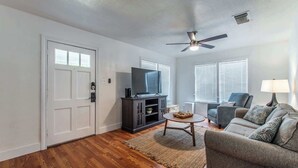 A living room with a dark wood floor, a gray sofa, a television on a cabinet, and a ceiling fan. - Texas Tranquility Retreat (Corpus Christi)