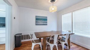 A dining room with a wooden table and white chairs, a painting on the wall, and a window with blinds. - Texas Tranquility Retreat (Corpus Christi)