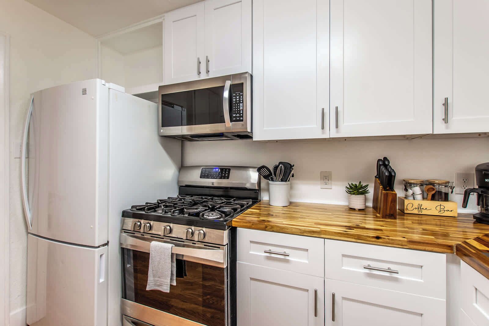 A kitchen with white cabinets, a stainless steel refrigerator, a microwave, a stove, and a coffee bar.