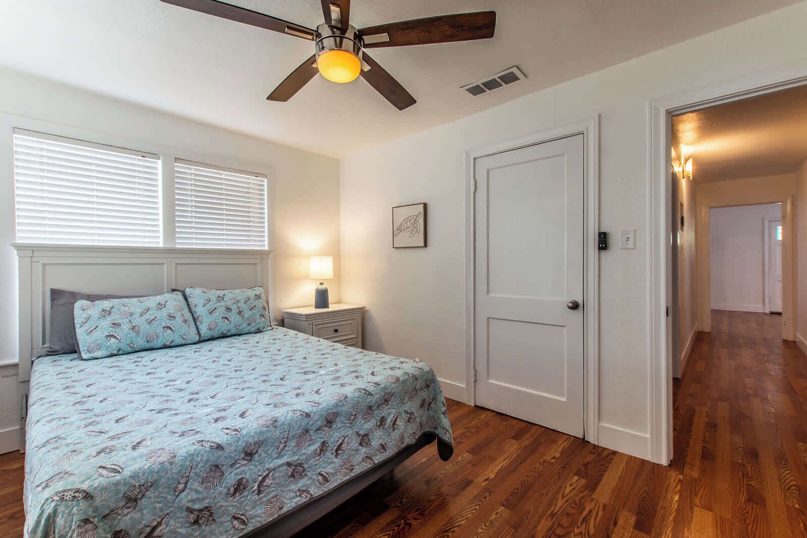 A bedroom with a bed, a nightstand, a ceiling fan, a door, and a hallway.