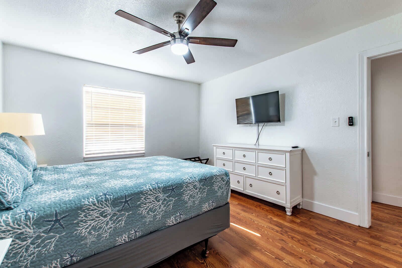 A bedroom with a bed, a dresser, a ceiling fan, a window with blinds, and a TV mounted on the wall.