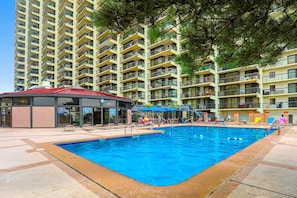 Ein Schwimmbecken mit einem Gebäude mit rotem Dach, umgeben von einem mehrstöckigen Haus mit Balkonen und blauem Himmel.
