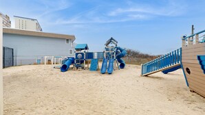 Un’area giochi sabbiosa con uno scivolo e strutture per arrampicarsi, adiacente a un edificio con tetto blu.