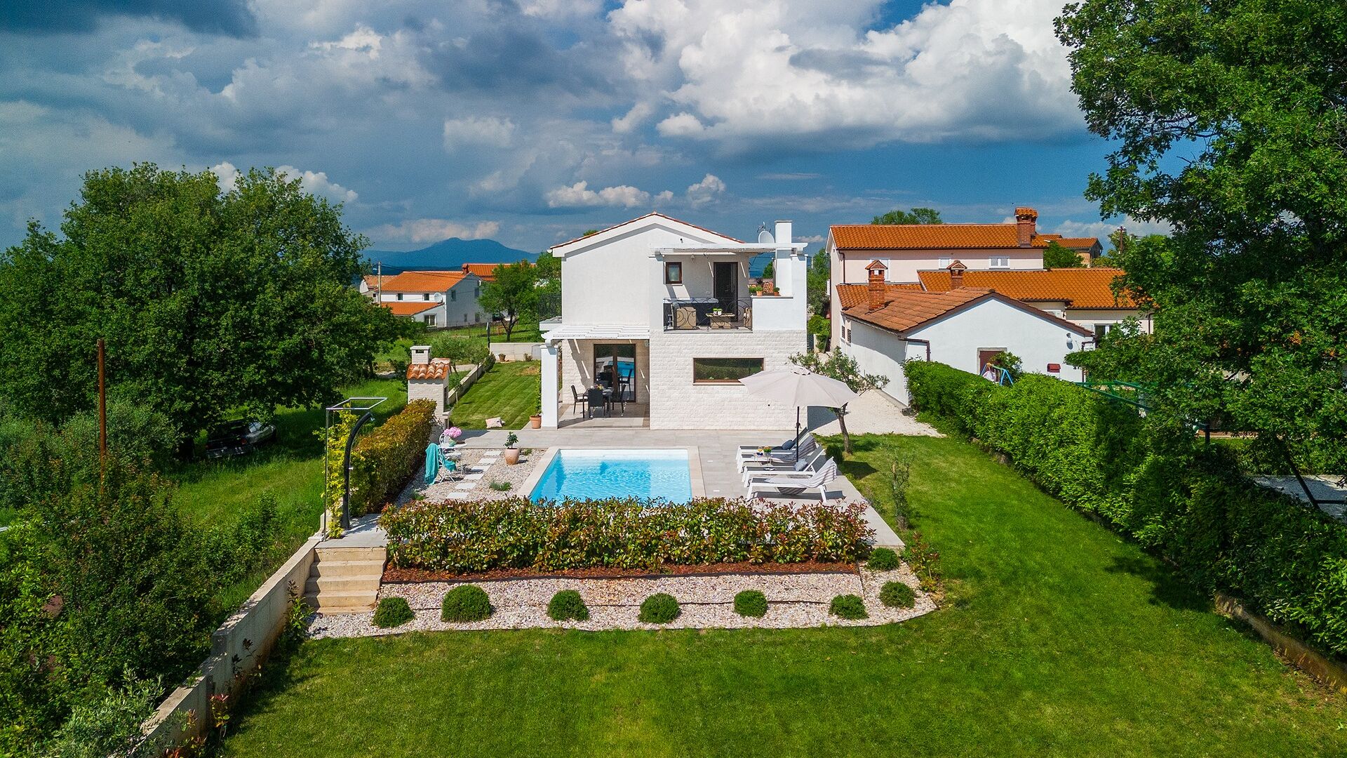 Una casa moderna de dos pisos amb piscina, envoltada de vegetació exuberant i un cel serè.