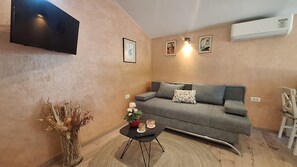 Ein Wohnzimmer mit grauem Sofa, einem kleinen schwarzen Couchtisch, einer Blumenvase und einem an der Wand befestigten Fernseher.