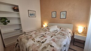 Ein Schlafzimmer mit einem Bett, Nachttischlampen, einem Nachttisch, einem Regal mit Büchern und zwei gerahmten Bildern an der Wand.