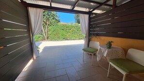 Eine überdachte Terrasse mit Tisch und Stühlen, einem Blumentopf und Blick auf einen Hecken- und Baumgarten.