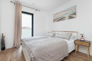 Ein Schlafzimmer mit Bett, Nachttisch, einem Fenster mit Vorhängen, einem Gemälde und einer Zimmerpflanze.