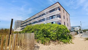 A multi-story building with balconies, a sandy beach in the foreground, and a clear sky. - Bimini 306 (Ocean City)