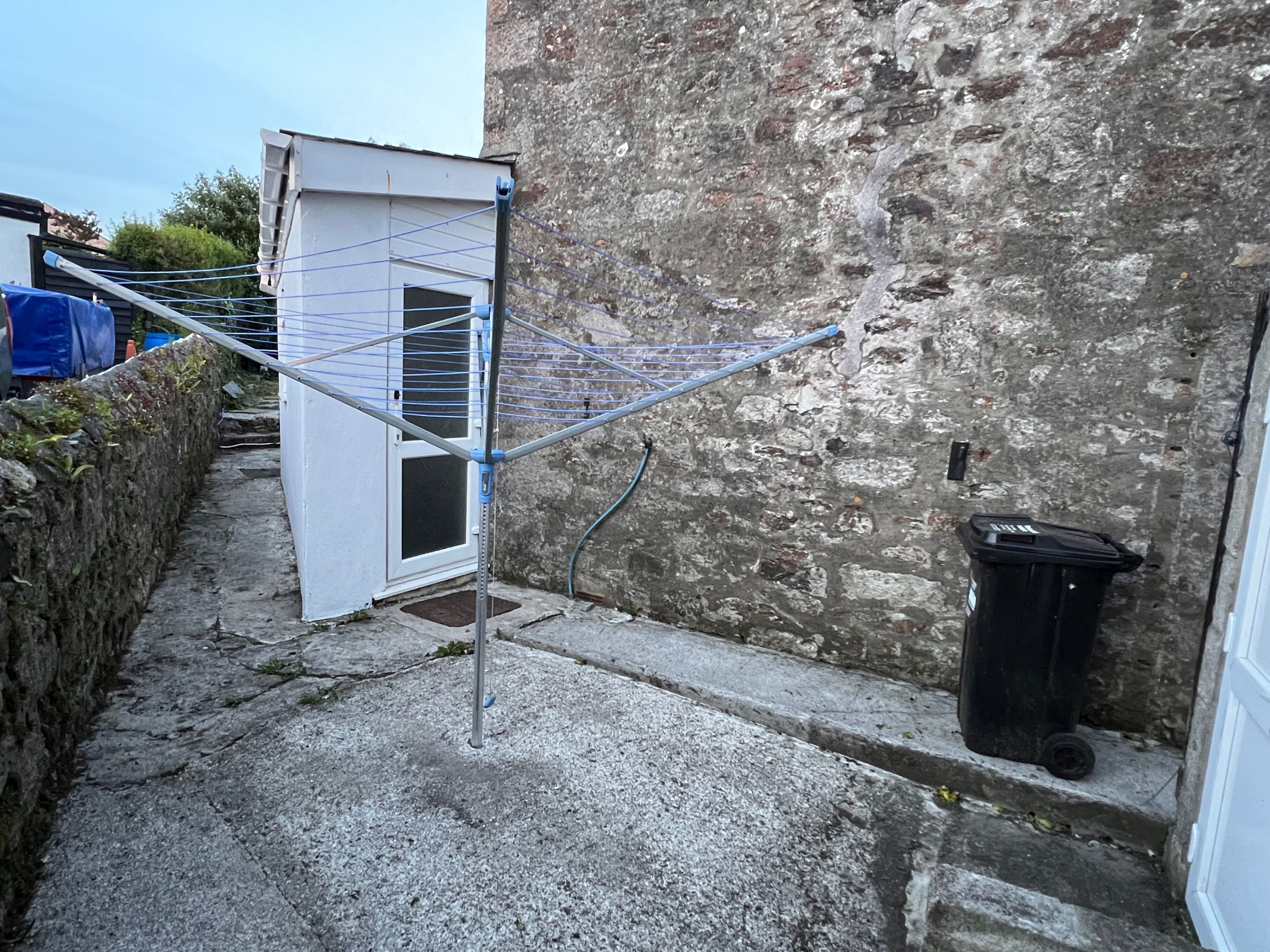 A narrow alley with a clothes drying rack, a small white building, and a black trash bin.