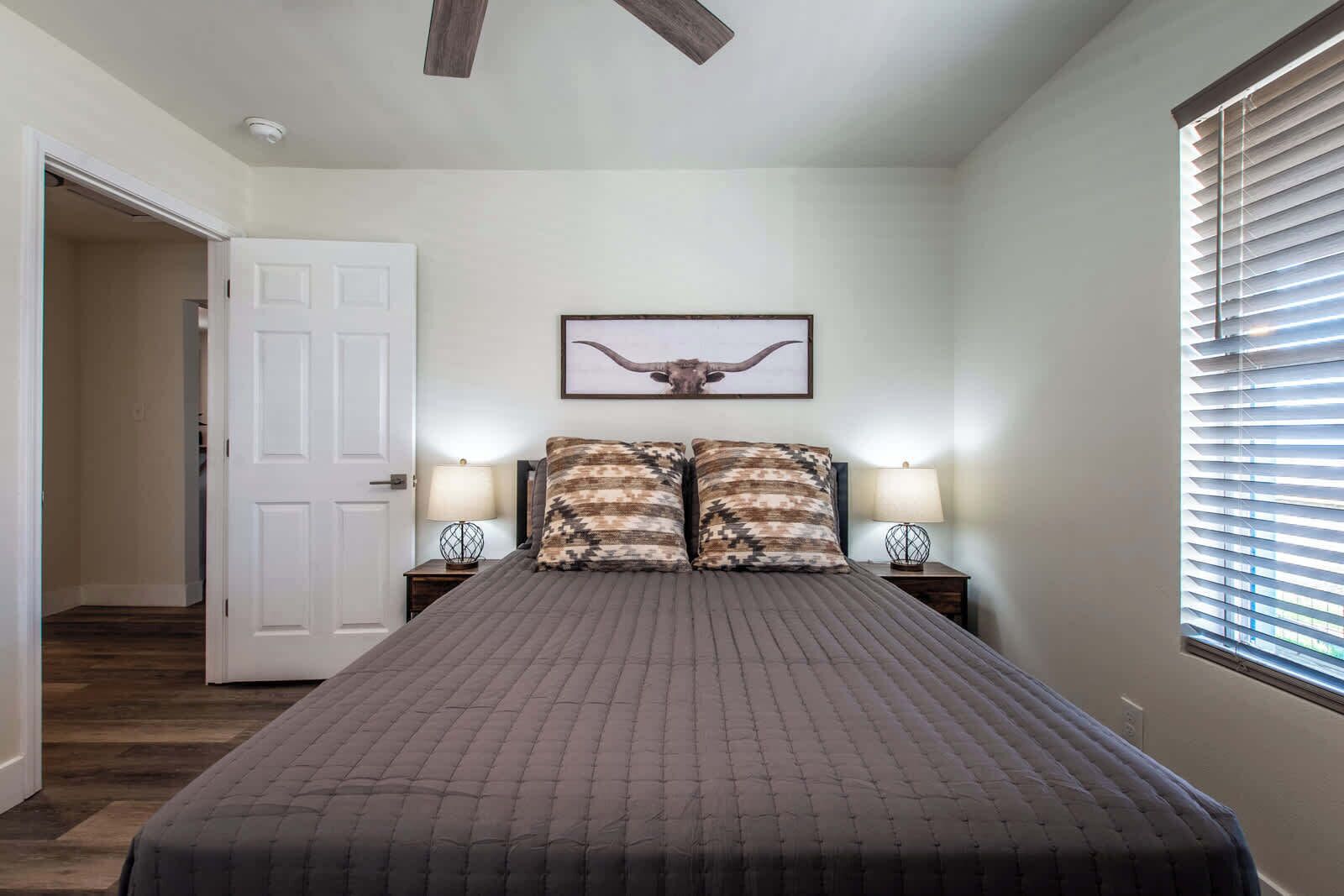 A bedroom with a bed, two nightstands, a ceiling fan, a door, and a window with blinds.