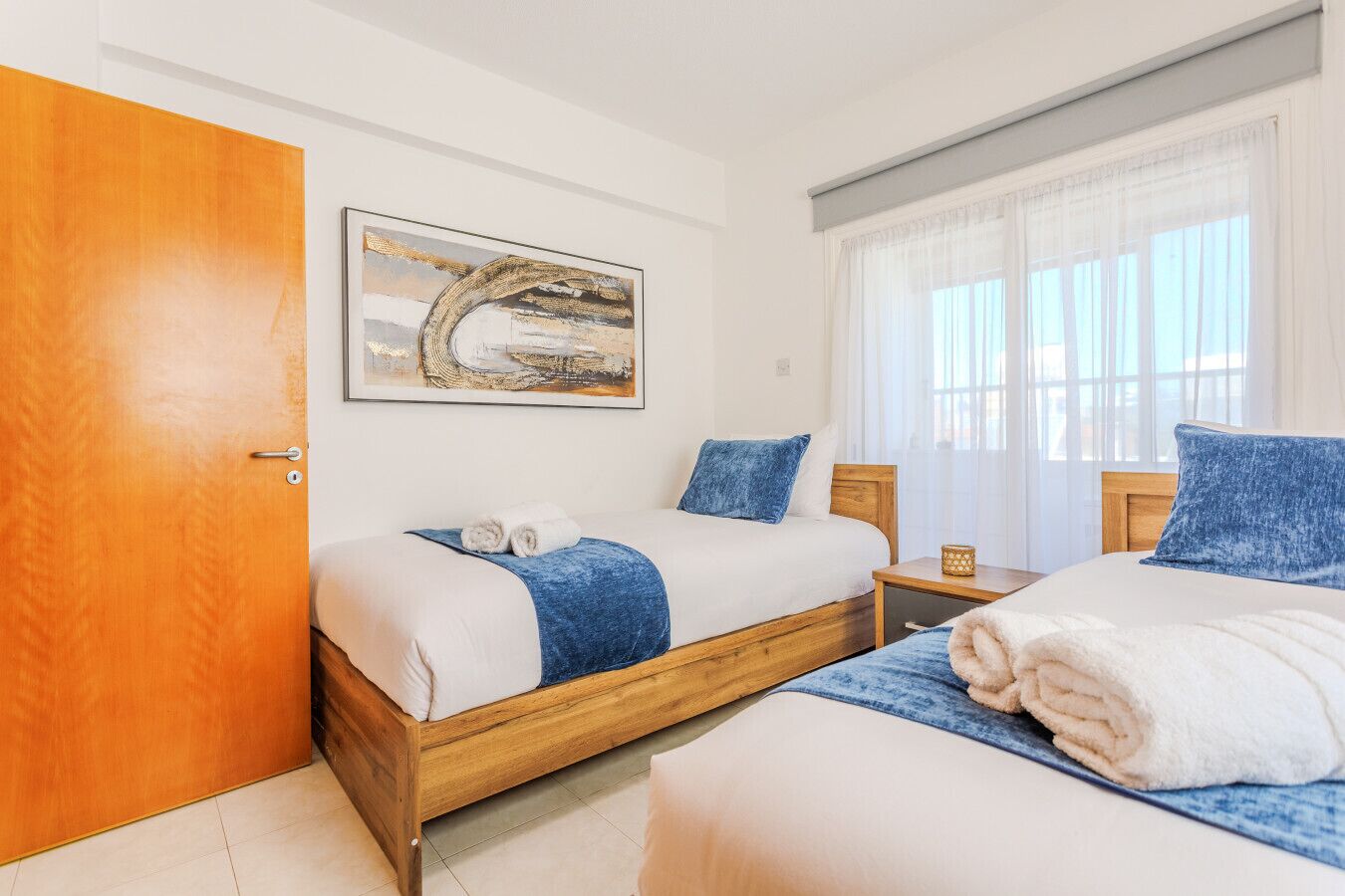Habitación de hotel con dos camas, una puerta de madera, un cuadro en la pared y una ventana con cortinas.