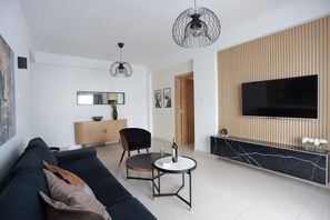 A modern living room with a black sofa, a round coffee table, a TV mounted on a wooden panel wall, and a pendant light. - TelMar Central Modern Retreat (Larnaka)