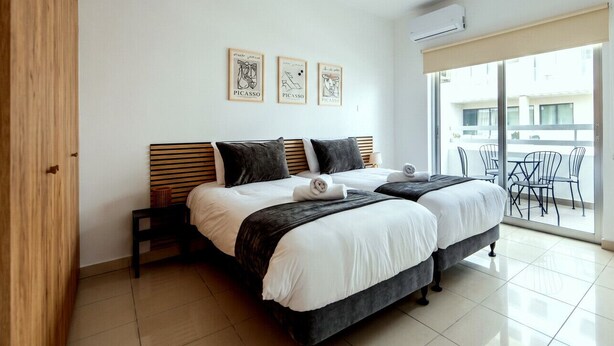 A double bed with a wooden headboard, two bedside tables, a large window with a view, and three framed artworks on the wall. - TelMar Central Modern Retreat (Larnaka)