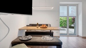 A modern dining area with a wooden table, a bowl of fruit, and a bottle. There is a bench with a fur throw, and a TV mounted on the wall. - Accessible Apartment '3' with Garden, Wi-Fi and Air Conditioning (Auer)
