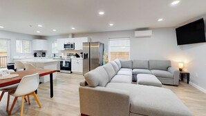 Una cocina moderna con armarios blancos, una mesa de comedor con sillas, un sofá seccional gris y un televisor de pantalla plana montado en la pared.