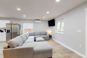 A modern living space with a sectional sofa, a flat-screen TV mounted on the wall, and a kitchen area with a refrigerator.