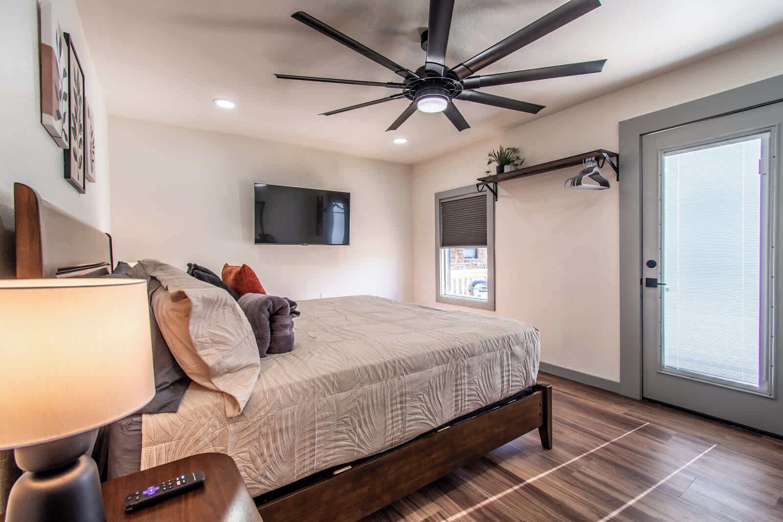 A bedroom with a bed, a ceiling fan, a TV mounted on the wall, a window with blinds, and a door with a window.