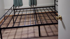A room with a metal bed frame, a window with curtains, a wooden floor, and a door with a brass doorknob. - 2-bedroom mobile home w/ AC at Elkwood Campground in Wolverine, MI with POOL! (Wolverine)