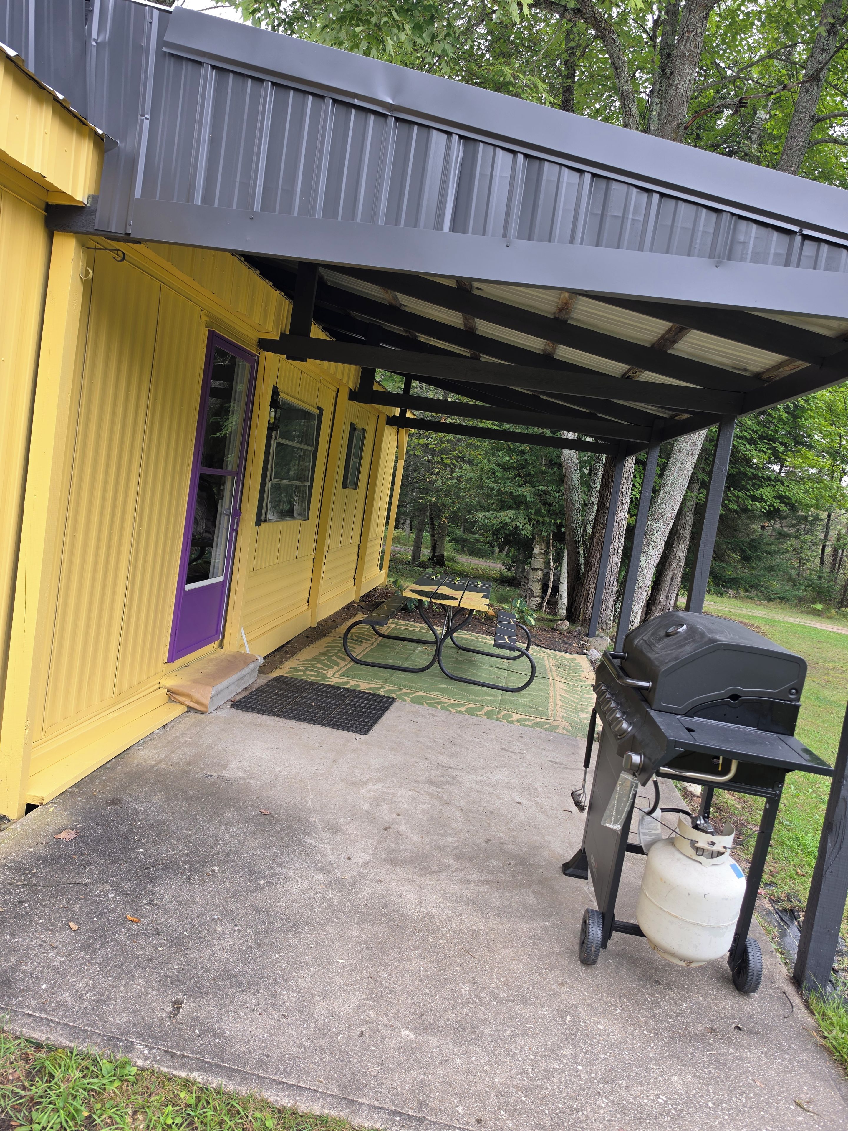 Un espacio exterior techado con una parrilla para barbacoa y tanque de propano, una mesa de picnic y un edificio amarillo con una puerta morada.
