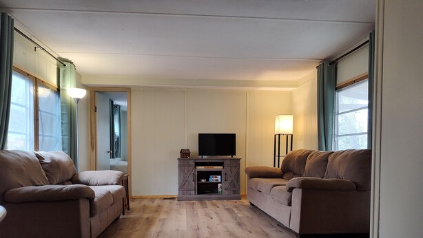 A living room with two sofas, a TV on a wooden cabinet, and a floor lamp. - 2-bedroom mobile home w/ AC at Elkwood Campground in Wolverine, MI with POOL! (Wolverine)