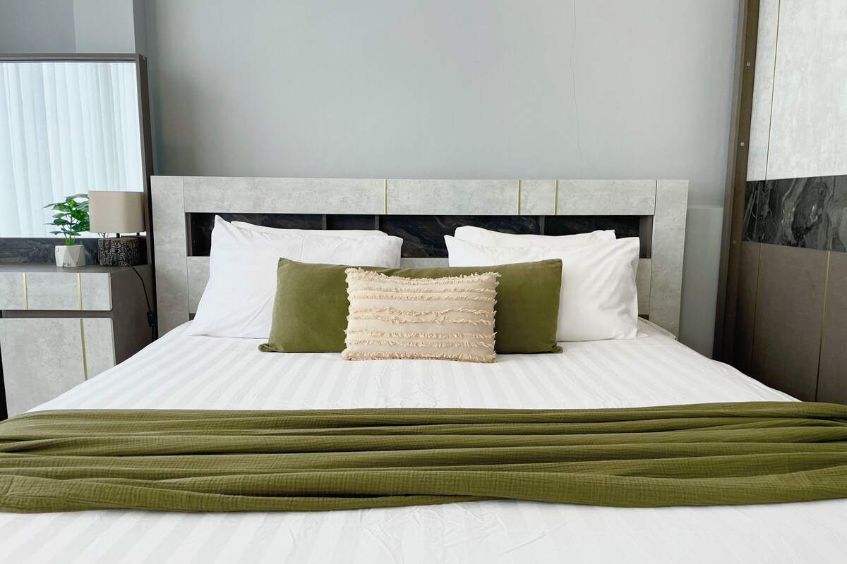 A neatly made bed with white linens and green accents, a headboard with a marble-like texture, and a bedside table with a lamp and a potted plant.