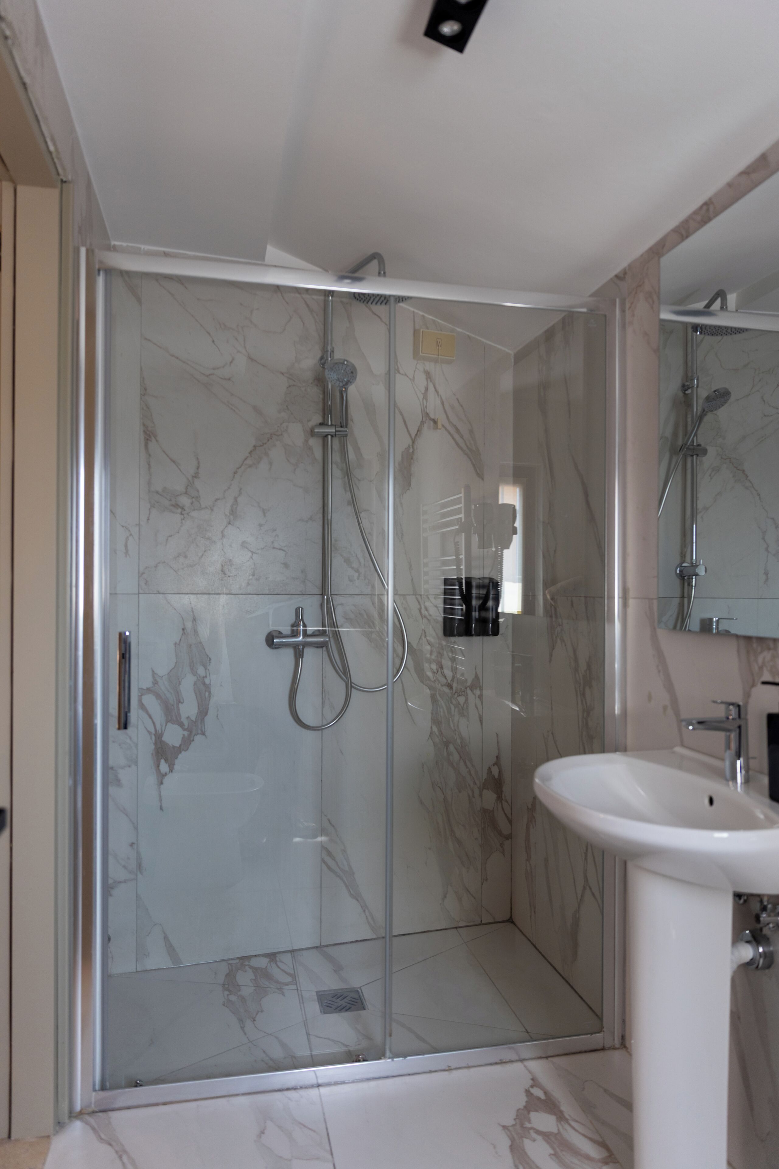Un baño moderno con ducha acristalada, paredes de mármol y un lavabo blanco.