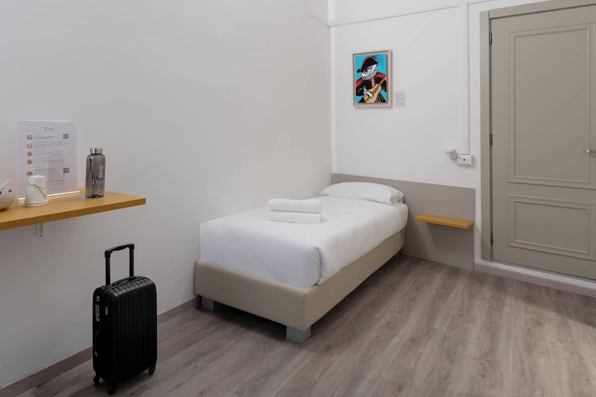 A hotel room with a bed, a small desk with a water bottle, a suitcase, and a painting on the wall.