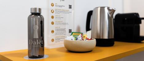 Een hotelkamer met een minibar, waterkoker, een schaal met snacks en een waterfles op een gele toonbank.