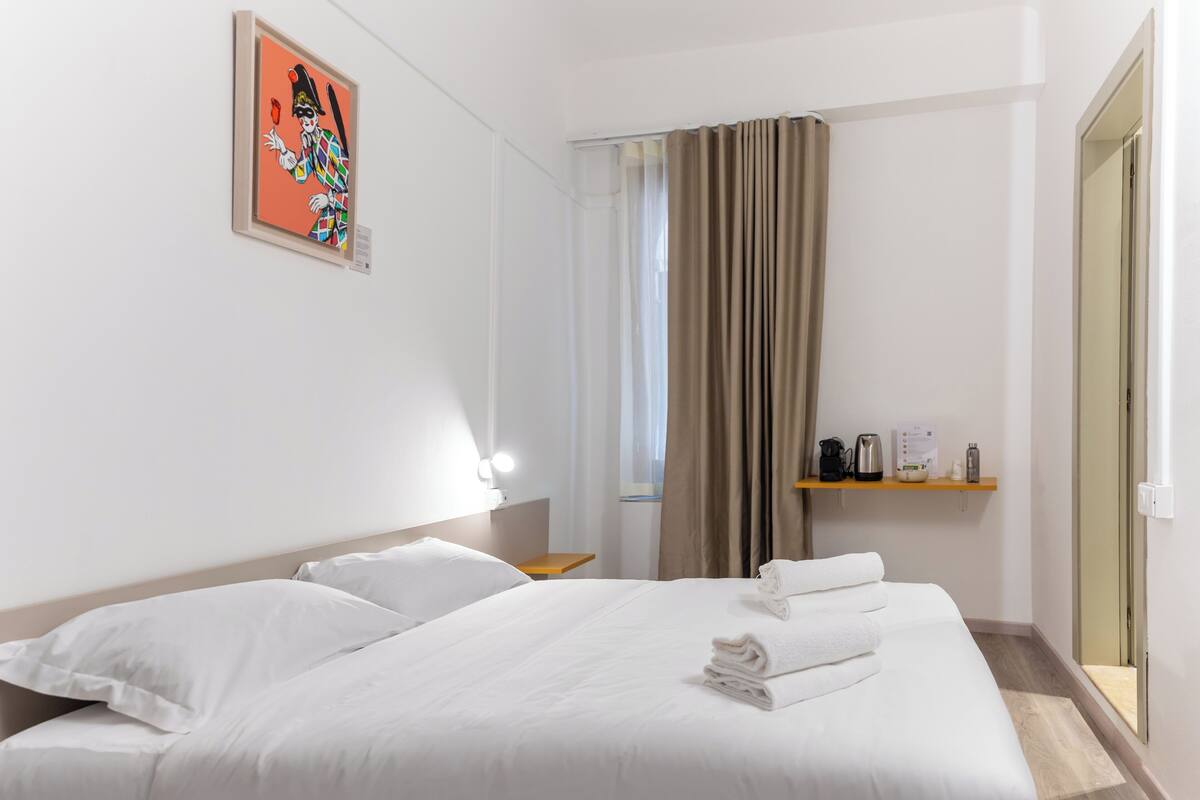 A hotel room with a large bed, a bedside table, a nightstand, a window with curtains, a wall-mounted light, and a small shelf with a kettle and other items.