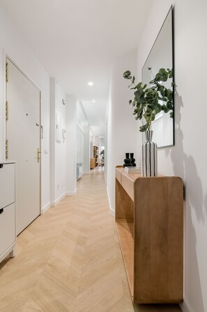 Ein Flur mit Holzboden, einer weißen Tür, einem Spiegel und einer Pflanze auf einer Holzkonsole.
