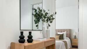 Une chambre moderne avec une console en bois, un miroir, une plante sur la table de chevet, un lit avec une couette blanche et une table de chevet avec une lampe.