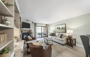 A living room with a sofa, coffee table, and TV. There are shelves with decorative items and a window with a view.