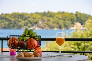 Et bord med en frugtkurv, et fad med bagværk, et glas appelsinjuice og et stearinlys, med udsigt over vand og grøn bevoksning.