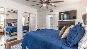 A bedroom with a large bed, a ceiling fan, a TV mounted above the bed, and a mirror on the wall. - Tranquil Terrace - Duplex #1 (Corpus Christi)