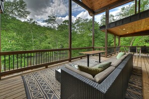 A covered outdoor deck with a wicker sofa, a table, and a view of a forested area.