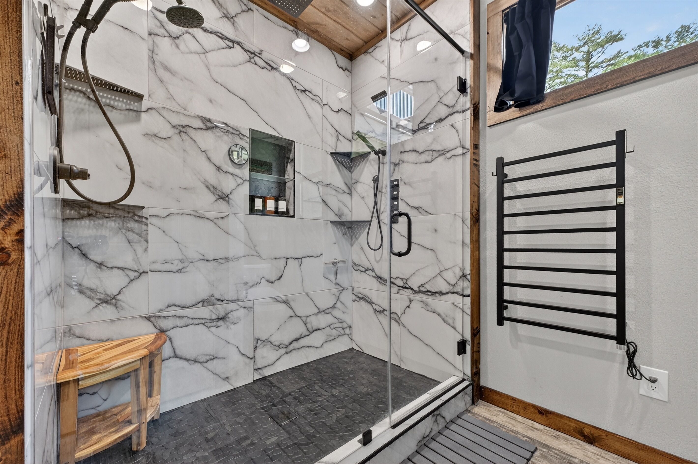 A modern bathroom with marble walls, a glass-enclosed shower, a wooden stool, and a towel rack.