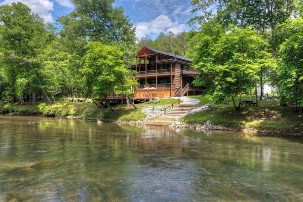 A wooden cabin with a deck, surrounded by trees and a lake.