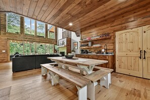 A wooden interior with a dining table, benches, a sofa, and a kitchen area with shelves and cabinets.